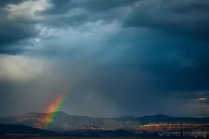 Fine art landscape photograph of a partial rainbow arc against desert landscape cliffs with dark clouds by Cramer Imaging