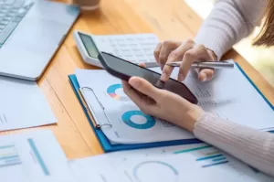 Photo of a woman holding a cell phone with a clipboard, calculator, and laptop surrounded by statistics paperwork