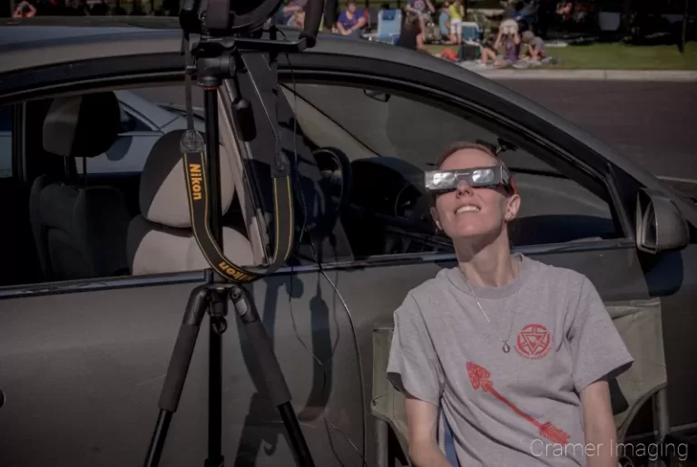 Photograph of landscape photographer Audrey Cramer wearing eclipse glasses and waiting with camera and car for the 2017 total solar eclipse