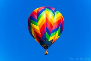 Fine art photograph of a rainbow-colored hot air balloons with chevron pattern against a blue sky by Cramer Imaging