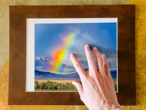 Photo of a hand adjusting the position of Cramer Imaging's landscape photo titled "Broken Rainbow" inside the window of a hinged brown mat