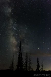 Cramer Imaging's fine art astrophotography photograph of the Milky Way in the night sky with clouds and trees in silhouette