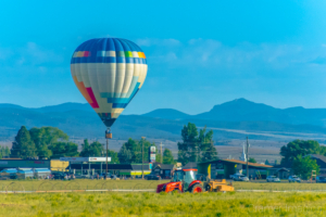 Cramer Imaging's fine art photo of a hot air balloon hovering over a farm field with tractor and a town in the background